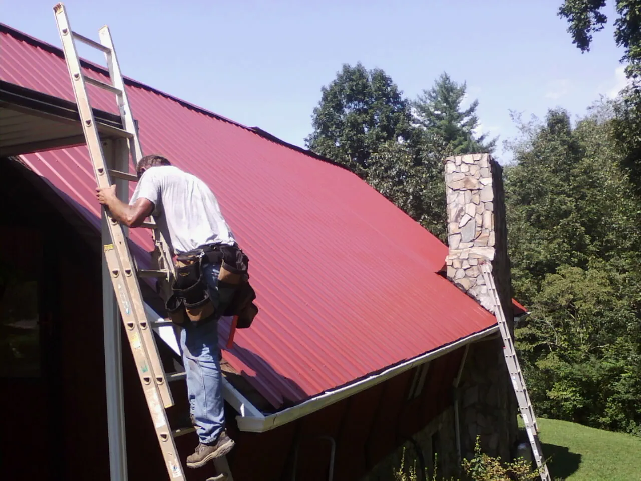 Roofer on a ladder beside a red metal roof and stone chimney — Soffit & Fascia Repair by Roofing Brothers in Douglassville
